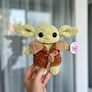 May include: A plush toy made of light green yarn with large black eyes and pink cheeks, wearing a brown textured vest. The creature has wide ears and a small round tag attached to its arm that features the text OVA TOYS in a circular design.