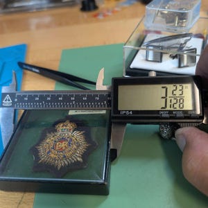 Puede incluir: Una insignia bordada del Royal Corps of Transport con una corona dorada y un emblema central sobre un fondo de tela oscura, dentro de una caja cuadrada. Un calibrador digital muestra una medida de 8,2 cm de ancho para el objeto.