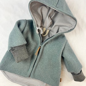 Op de afbeelding: Een tealkleurige jas met capuchon en een grijs-witte gestreepte voering. De jas heeft een ritssluiting, grijze manchetten en een klein bruin label. De capuchon is gevoerd met een grijs-witte gestreepte stof.