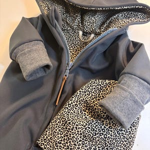 Könnte beinhalten: Grauer Kapuzenoverall mit Leopardenmuster-Futter und passender Mütze. Der Overall hat einen Reißverschluss und einen grauen Besatz an Kapuze und Ärmelbündchen.