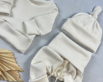 May include: A three-piece baby outfit in a cream color. The outfit includes a long-sleeved shirt, pants, and a knotted beanie.