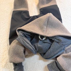 Può includere: Una tuta con cappuccio grigia e beige con chiusura a zip. La tuta presenta un corpo grigio scuro con dettagli beige e grigi sul cappuccio, sulle maniche e sulle gambe. L'indumento è realizzato in un materiale morbido e strutturato.