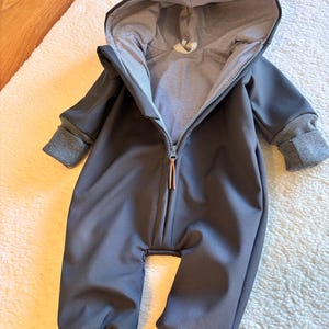 Op de afbeelding: Een donkergrijze baby-overall met een capuchon en een lange ritssluiting. De binnenkant van de capuchon en de manchetten zijn gevoerd met een grijs-witte gestreepte stof. De manchetten en enkels zijn donkerder grijs.