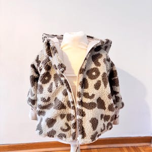 Könnte beinhalten: Kapuzenjacke für Kinder mit Leopardenmuster in Grau- und Brauntönen. Die Jacke hat einen durchgehenden Reißverschluss und wird auf einer weißen Schaufensterpuppe präsentiert. Das Innenfutter der Jacke ist hellbeige.