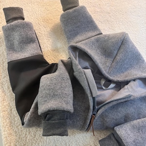 Könnte beinhalten: Grau-schwarzer Overall mit Kapuze und durchgehendem Reißverschluss. Die Manschetten und das Kapuzenfutter sind dunkler grau. Der Overall ist aus einem weichen, warmen Material gefertigt, ideal für kühleres Wetter.