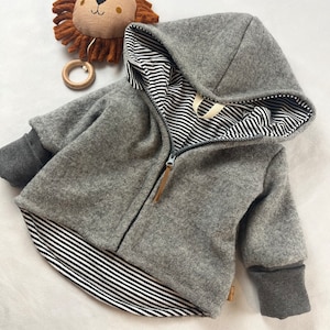 Könnte beinhalten: Eine graue Kapuzenjacke mit einem schwarz-weiß gestreiften Futter. Die Jacke hat einen Reißverschluss und einen braunen Zipper. Die Ärmel haben graue Bündchen.