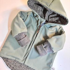 Könnte beinhalten: Eine salbeigrüne Kapuzenjacke mit einem schwarzen und weißen Leopardenmusterfutter. Die Jacke hat einen Reißverschluss und graue gerippte Bündchen.