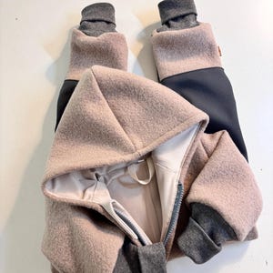 Könnte beinhalten: Ein Overall mit Kapuze aus strukturiertem, altrosa wollartigem Stoff mit dunkelgrauen Rippbündchen an Ärmeln und Beinen. Das Kleidungsstück verfügt über einen Frontreißverschluss, ein helles Innenfutter und ein kleines braunes Etikett an der Seite.