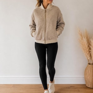 Puede incluir: Chaqueta bomber beige con cremallera y ribetes color crema, combinada con leggings negros y zapatillas blancas. La chaqueta tiene un aspecto suave y texturizado. El conjunto es informal y cómodo.