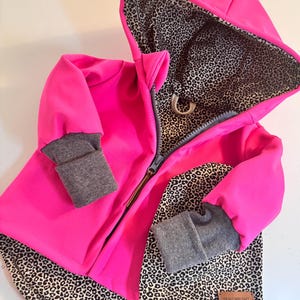 Könnte beinhalten: Eine rosa Kapuzenjacke mit Leopardenmuster-Futter und passender Mütze. Die Jacke hat einen Reißverschluss und graue Bündchen. Die Mütze hat ein braunes Leder-Etikett mit dem Text "Frau Wipunkt".