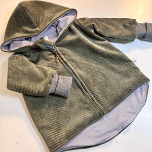 Könnte beinhalten: Olivgrüne Cord-Kapuzenjacke mit durchgehendem Reißverschluss. Die Jacke hat graue Rippbündchen und ein gestreiftes Futter. Die Kapuze ist mit dem gleichen gestreiften Stoff gefüttert. Ein kleiner Lederzug ist am Reißverschluss befestigt.