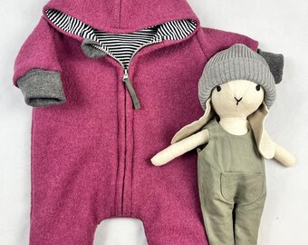 Peut inclure: Une combinaison rose à capuche avec des bordures grises et une fermeture éclair. La combinaison est faite d'un tissu doux et duveteux. Un lapin en peluche gris et blanc est assis à côté de la combinaison.
