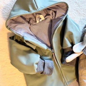 Può includere: Giacca con cappuccio verde chiaro con chiusura a zip completa. La giacca ha polsini a coste grigi e fodera a righe. Il cappuccio è completamente foderato con lo stesso tessuto a righe. La giacca è in materiale morbido.