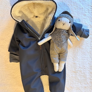 Op de afbeelding: Een donkergrijze baby-sneeuwoverall met een capuchon gevoerd met crèmekleurig imitatiebont. De sneeuwoverall heeft een ritssluiting en grijze manchetten. Een knuffelkonijn met een patroonhoed en sjaal ligt naast de overall.