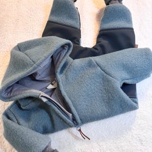 Könnte beinhalten: Hellblauer Baby-Schneeanzug mit Kapuze und durchgehendem Reißverschluss. Die Bündchen und das Kapuzenfutter sind grau. Der Schneeanzug hat ein weiches, strukturiertes Aussehen und ist für Wärme und Komfort konzipiert.