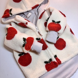 Könnte beinhalten: Eine weiße Kapuzenjacke mit rotem Apfelmuster. Die Jacke hat einen Reißverschluss und ein graues Innenfutter. Die Ärmelbündchen und die Kapuze sind rot eingefasst. Kinderjacke.