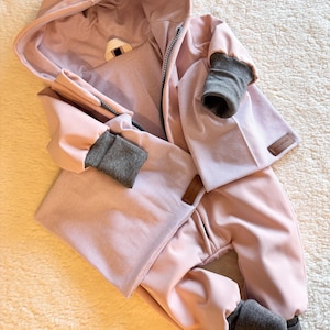 Könnte beinhalten: Hellrosa Baby-Schneeanzug mit Kapuze und Reißverschluss. Die Manschetten und das Innenfutter der Kapuze sind grau. Der Stoff hat eine dezente Streifenstruktur. Der Schneeanzug ist für Wärme und Komfort konzipiert.