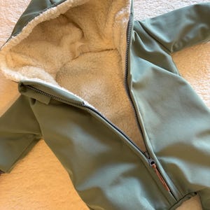 Op de afbeelding: Olijfgroene baby-sneeuwoverall met capuchon en een lange ritssluiting. De binnenkant is gevoerd met een zachte, crèmekleurige fleece. De sneeuwoverall heeft lange mouwen en bedekt de benen, ontworpen voor koude weersomstandigheden.