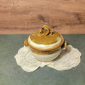 May include: A round ceramic lidded bowl with two side handles and a loop handle on the lid. The bowl features a two-tone glaze with a speckled mustard yellow and brown top section transitioning into a smooth, creamy off-white base color.