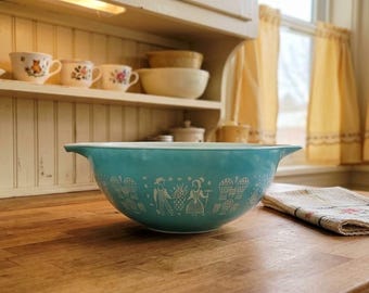 May include: A turquoise mixing bowl with a white pattern featuring figures and fruit designs. The bowl has two side handles and a smooth, glossy finish. It is positioned on a wooden surface, showcasing a classic kitchen aesthetic with a wide rim.