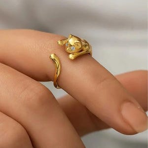 Op de afbeelding: Een goudkleurige open ring met een sculpturaal kattenontwerp. De kattenfiguur bevindt zich aan één uiteinde van de band, waarbij de kop en voorpoten op de vinger rusten. De ogen van de kat zijn geaccentueerd met twee kleine, heldere, fonkelende steentjes.