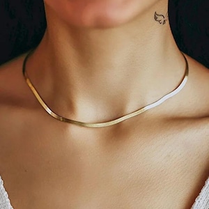 Puede incluir: Un collar de cadena tipo espiga en tono dorado con un diseño de eslabones planos, flexibles y lisos que descansa cerca de la base del cuello. El collar tiene un acabado metálico pulido que refleja la luz en toda su superficie.