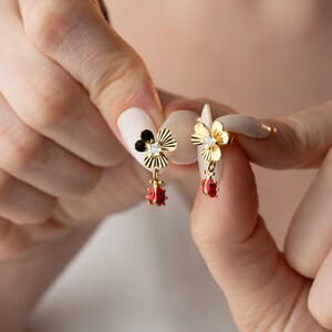 Pode incluir: Um par de brincos dourados com um pino em forma de flor com uma pedra transparente central e um pingente de joaninha vermelho e preto pendurado. As pétalas da flor têm uma textura estriada e a joaninha apresenta pontos pretos detalhados.