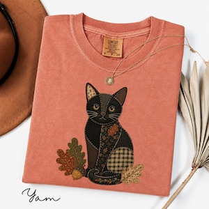 Puede incluir: Una camiseta de manga corta color terracota con un gráfico central bordado de un gato negro compuesto por varios patrones de tela, incluyendo cuadros y flores. El gato está rodeado de hojas de roble bordadas y una bellota.