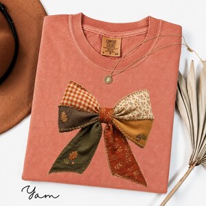 Könnte beinhalten: Ein terrakottafarbenes T-Shirt mit einer großen, mittigen Grafik einer Schleife im Patchwork-Stil. Die Schleife besteht aus verschiedenen Stoffmustern, darunter orangefarbenes Vichy-Karo, Blumenmuster und erdige Farbtöne, akzentuiert durch kleine gestickte Eichenblätter.