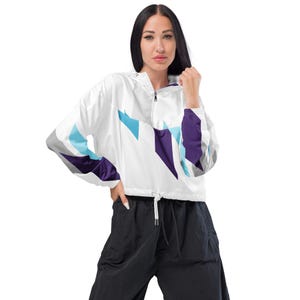 May include: A white cropped windbreaker jacket featuring a quarter-zip neckline, a drawstring hem, and long sleeves with a geometric pattern in shades of purple, light blue, and gray. The jacket is paired with loose-fitting black trousers.