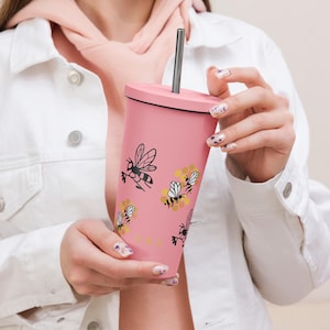 Puede incluir: Un vaso rosa con tapa y pajita de metal, que presenta un patrón repetitivo de abejas dibujadas con líneas negras y formas de panal amarillas. El vaso tiene un acabado mate suave y una forma cilíndrica.