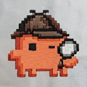 Può includere: Una toppa ricamata raffigurante una creatura arancione pixelata che indossa un cappello marrone. La creatura ha un occhio nero quadrato, una piccola bocca nera e tiene in mano una lente d'ingrandimento circolare con lente bianca e cornice nera.