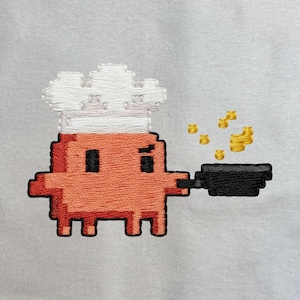 以下が含まれることがあります： 明るいグレーの布地を背景にした、刺繍によるピクセルアートのキャラクター。キャラクターはオレンジ色で白いシェフハットを被り、黒いフライパンを持っています。フライパンの上には、湯気や食材を表す小さな黄色い四角いピクセルが浮かんでいます。