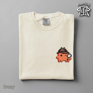 Puede incluir: Una camiseta de color marfil con un pequeño bordado de pixel art en el pecho. El diseño muestra un personaje naranja con un sombrero de detective marrón que sostiene una lupa. La camiseta tiene cuello redondo y una etiqueta de la marca Comfort Colors.