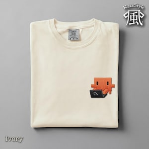 Puede incluir: Una camiseta de cuello redondo color marfil con un pequeño bordado en la parte inferior derecha. El diseño muestra un personaje naranja pixelado sentado frente a una computadora portátil negra pixelada. La camiseta tiene una etiqueta Comfort Colors.