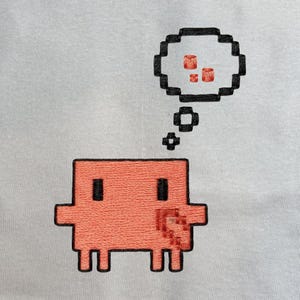 Puede incluir: Una tela gris claro con un personaje pixelado bordado en color naranja coral, con ojos negros y un pequeño detalle de corazón. Sobre el personaje hay una burbuja de pensamiento pixelada negra con tres pequeños cilindros naranja coral.