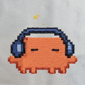 Puede incluir: Un diseño bordado de pixel art sobre una tela gris claro. El diseño presenta un personaje rectangular naranja con dos líneas negras horizontales como ojos, usando auriculares azul oscuro, con una pequeña forma pixelada amarilla arriba.