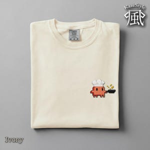 Könnte beinhalten: Ein elfenbeinfarbenes T-Shirt mit Rundhalsausschnitt und einer kleinen Pixel-Art-Stickerei auf der Brust. Das Motiv zeigt eine rote, quadratische Figur mit weißer Kochmütze, die eine schwarze Pfanne mit kleinen gelben Lebensmitteln hält.