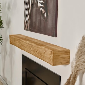 May include: A rectangular wooden mantel shelf with a natural light brown wood grain finish and visible knots, mounted horizontally on a white wall above a black fireplace insert, positioned below a framed zebra print wall art piece.