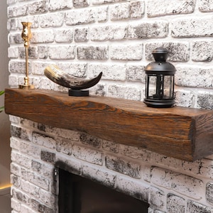 May include: A rectangular, dark brown wooden fireplace mantel with a prominent wood grain texture. The mantel is mounted on a white brick wall and holds a gold-colored candle holder, a curved animal horn on a black base, and a black metal lantern.