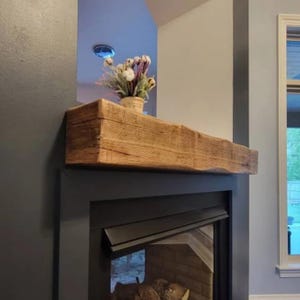 May include: A rectangular fireplace mantel made of natural, unfinished wood with a prominent grain pattern. The mantel is mounted horizontally above a black fireplace insert. A small ceramic vase containing dried flowers sits on top of the wood.