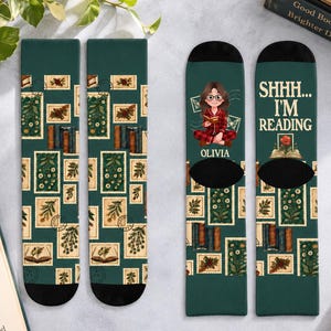 May include: A pair of dark green crew socks featuring a repeating pattern of vintage-style postage stamps with botanical and book illustrations. One sock displays a cartoon character reading a book with the text "SHHH... I'M READING" and "OLIVIA".