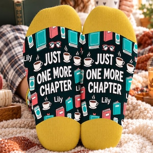 Op de afbeelding: Een paar zwarte sokken met gele hielen en tenen, voorzien van een herhalend patroon van boeken, koffiekopjes en brillen. De tekst "JUST ONE MORE CHAPTER" is in het wit gedrukt, gevolgd door de naam "Lily" in een kleiner wit lettertype.