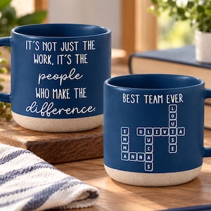 May include: Two navy blue ceramic mugs with a speckled beige base. The larger mug features the white text: IT'S NOT JUST THE WORK, IT'S THE people WHO MAKE THE difference. The smaller mug displays BEST TEAM EVER and a crossword puzzle design.