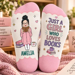 May include: A pair of white and pink socks featuring a cartoon illustration of a person with glasses reading a book, the name AMELIA, and the text JUST A GIRL WHO LOVES BOOKS, accompanied by small heart and star graphics on the fabric surface.