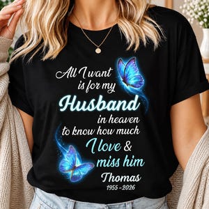 May include: A black short-sleeved t-shirt featuring two glowing blue butterflies and white text that reads: All I want is for my Husband in heaven to know how much I love & miss him Thomas 1955-2026. The shirt has a crew neckline.