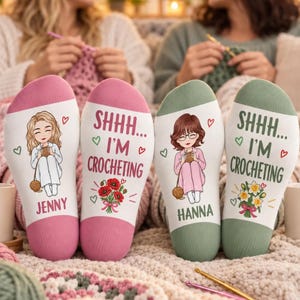 May include: Two pairs of white socks with coloured heels and toes. One pair features a blonde character, the name Jenny, and red poppies. The other pair features a brunette character, the name Hanna, and yellow daffodils. Both pairs display the text SHHH... I'M CROCHETING.
