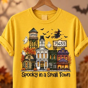 May include: A mustard yellow t-shirt featuring a graphic of a small town scene with a water tower labeled CARTHAGE, a sign reading 75633, ghosts, a witch on a broomstick, bats, and pumpkins. The text Spooky in a Small Town is printed below.