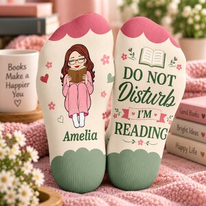May include: A pair of cream-coloured socks with scalloped pink and green cuffs. The left sock features a cartoon of a person with brown hair and glasses reading a book, with the name Amelia below. The right sock displays the text DO NOT DISTURB I'M READING with a book icon and floral accents.