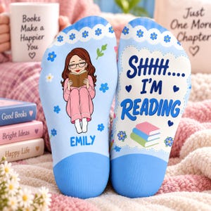 May include: A pair of light blue socks featuring a white scalloped trim with blue flowers. One sock displays a cartoon of a person with brown hair and glasses reading a book, with the name EMILY below. The other sock says SHHH.... I'M READING.